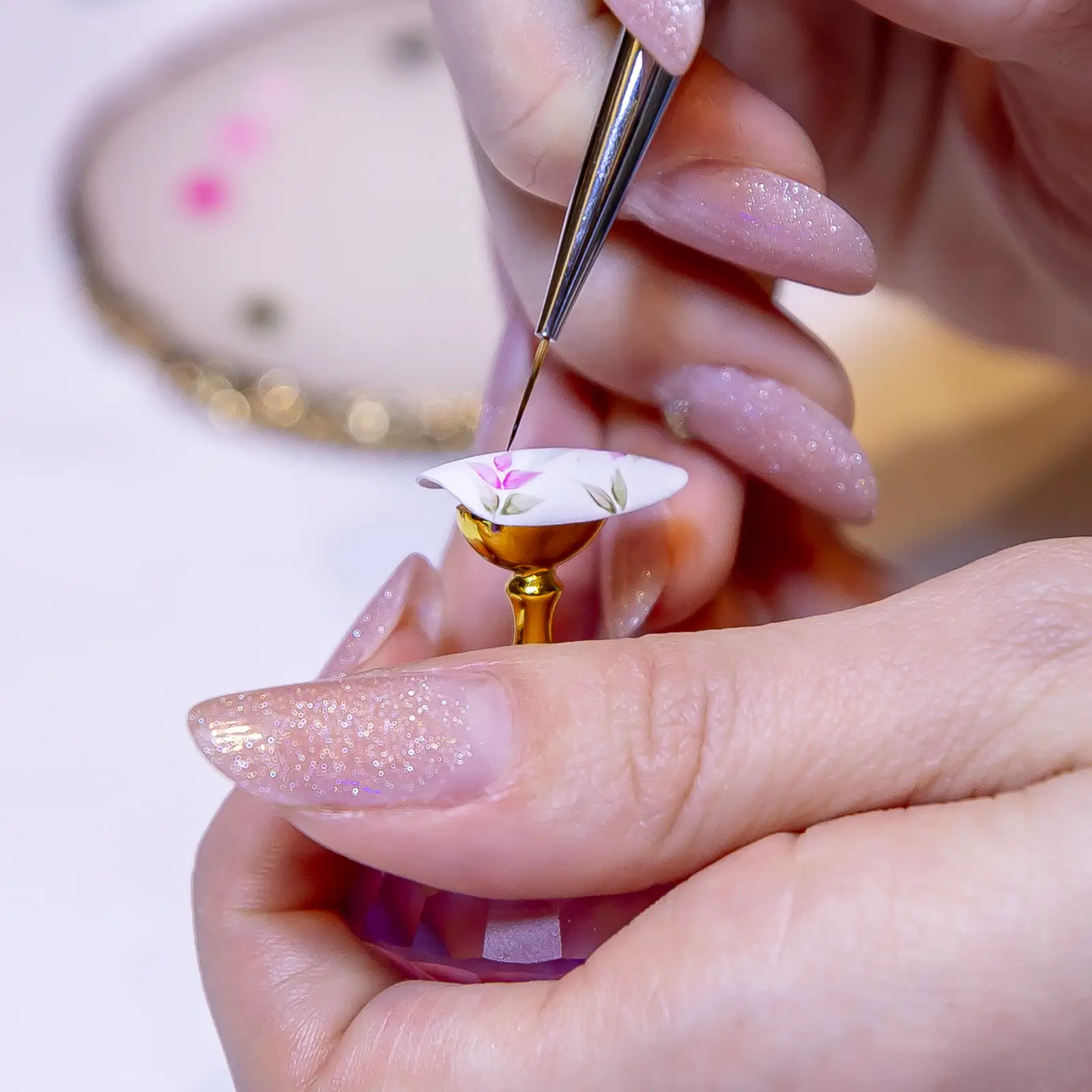 A fine brush painting pink leaves onto a white nail tip held on a gold stand